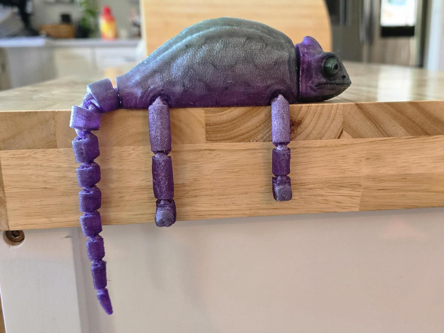 Articulated rubber chameleon with legs and tail hanging off of the side of a butcher block